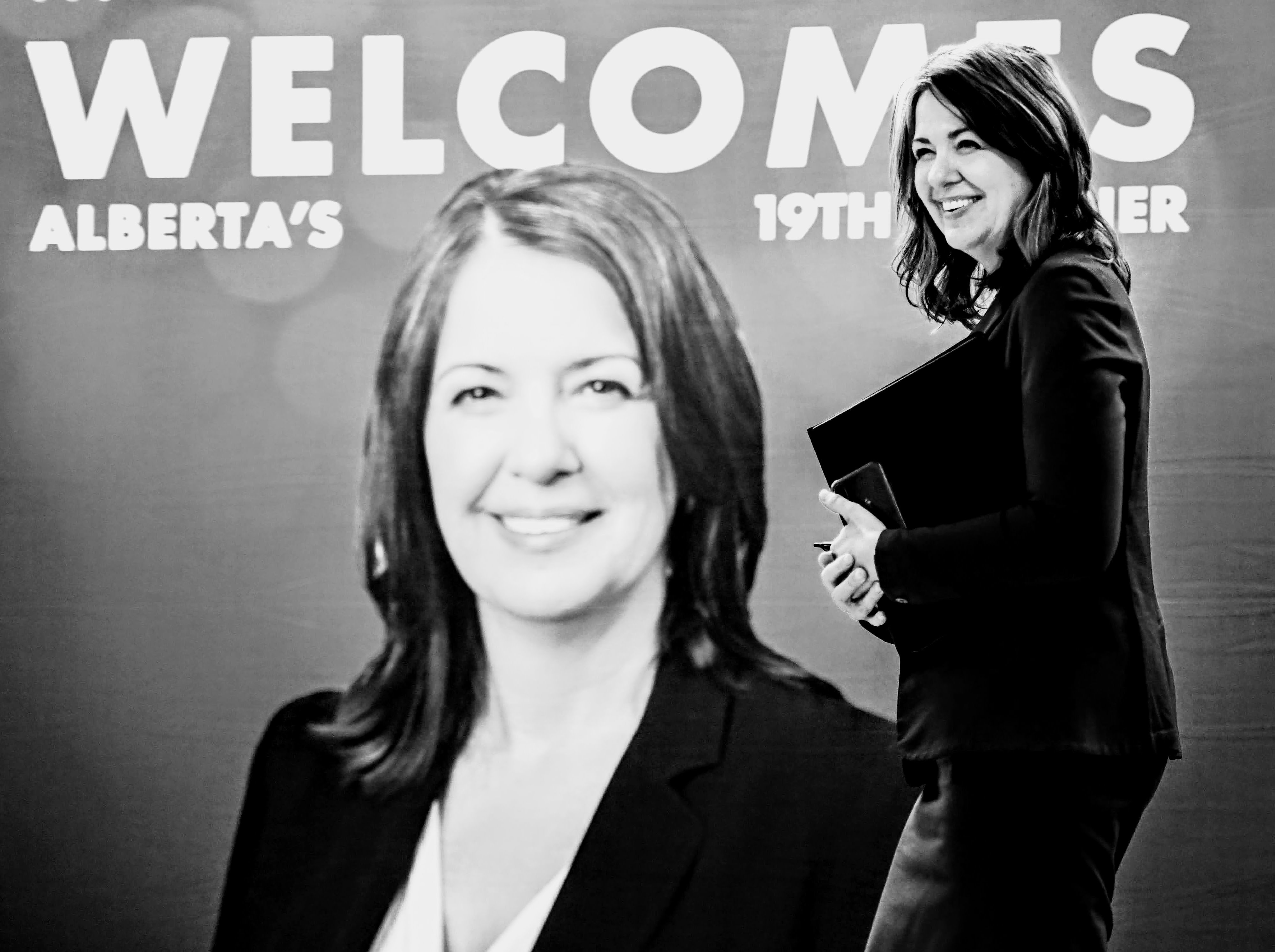 A black and white image shows two photos of the same smiling 50-ish light-skinned woman wearing a dark suit and holding a binder and phone.