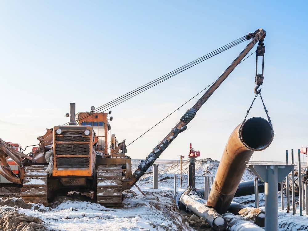 A large bulldozer with a boom arm holds a section of pipeline. The sky is clear and there is snow on the ground.