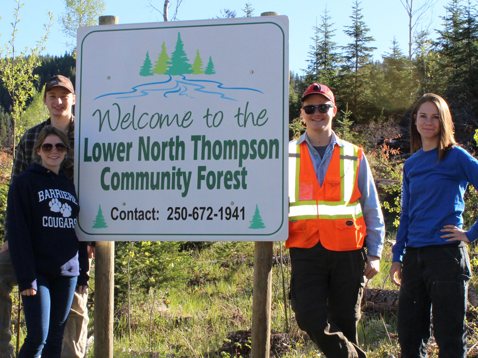 Four people stand beside a sign that says 'Welcome to the Lower North Thompson Community Forest,' with trees in the background.