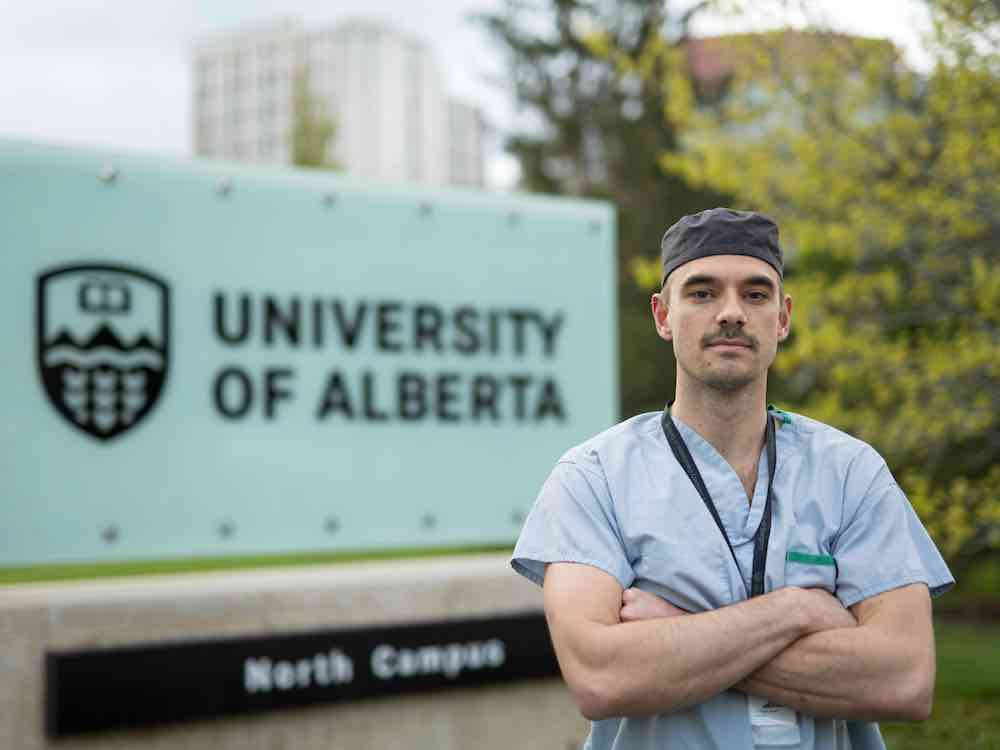 Dunavan Morris-Janzen is wearing light-blue medical scrubs and a dark grey surgical cap. He has light skin and stands with his arms crossed in front of a sign that reads 'University of Alberta.'