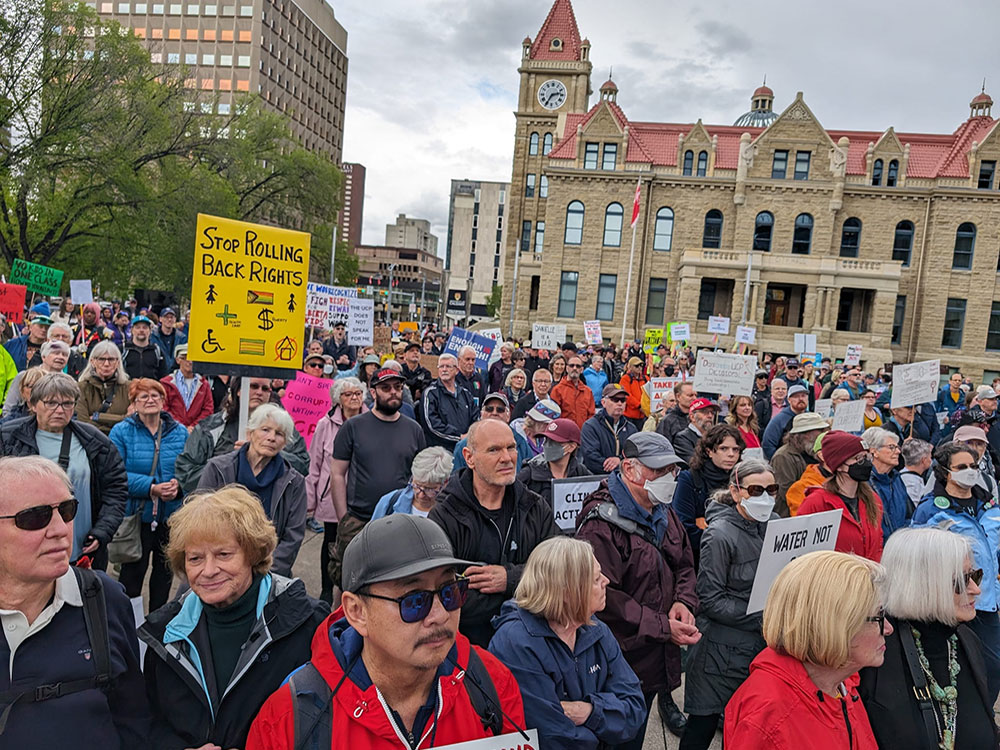 A large crowd of people stand outdoors. A sandstone building with a clock tower is in the background.