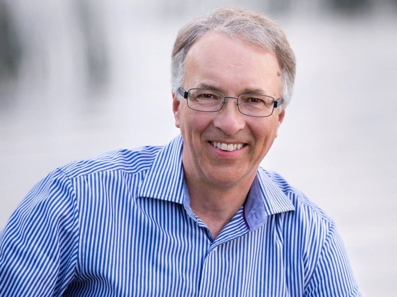 A light-skinned man with glasses and short grey hair smiles at the camera. He wears a blue and white striped shirt.
