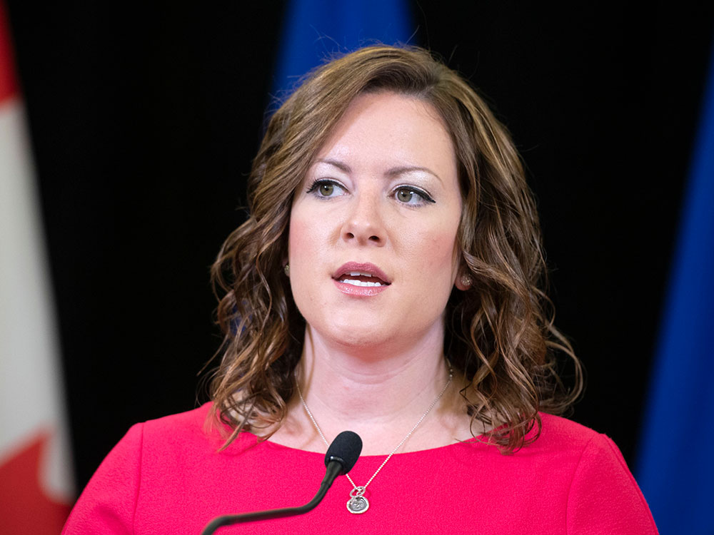A 39-year-old light-skinned woman with brown hair stands a podium wearing a red shirt.