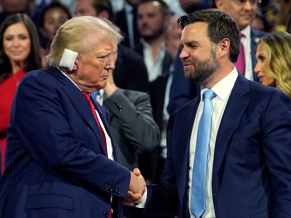 An older man in a blue suit with bandaged ear shakes hand with a younger man in blue suit and with a trimmed beard.
