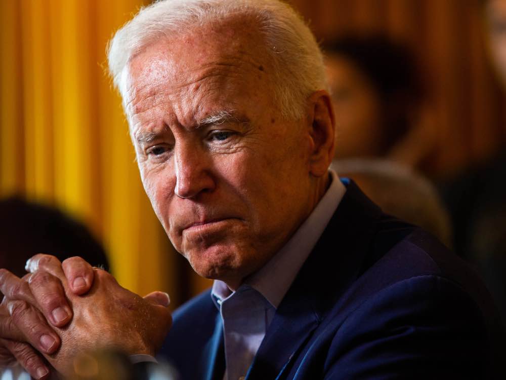 An 80-ish man with a ruddy complexion sits with hands clasped. His face is lined and wrinkled.