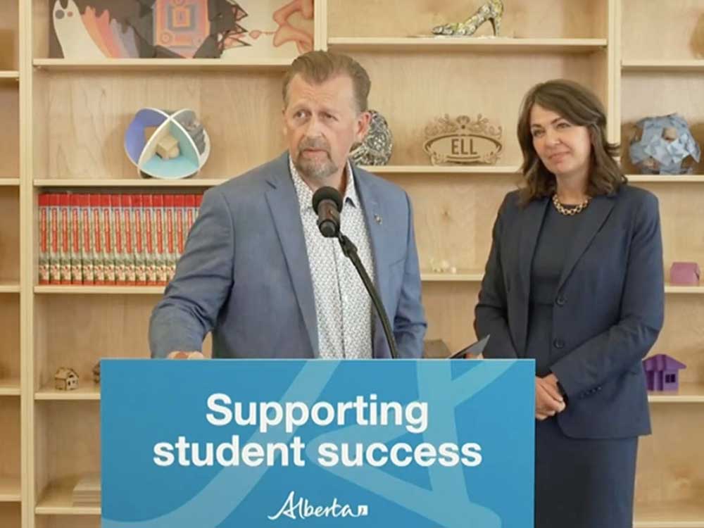 A middle-aged man with light skin and a beard, wearing casual clothes, stands at a podium. Behind him, a 50-ish woman wearing a dark suit, stands with her hands clasped.