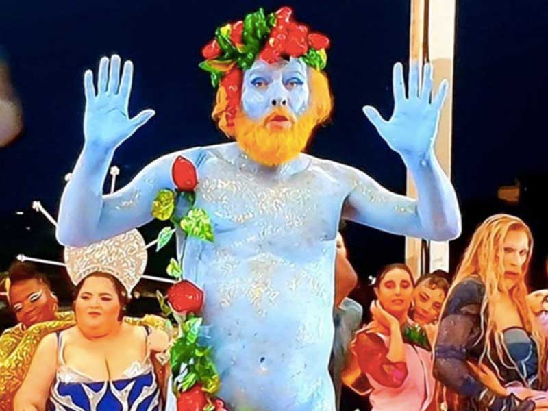 A man painted blue with a yellow beard and fruit crown sprawls on a table, with odd-looking characters behind.