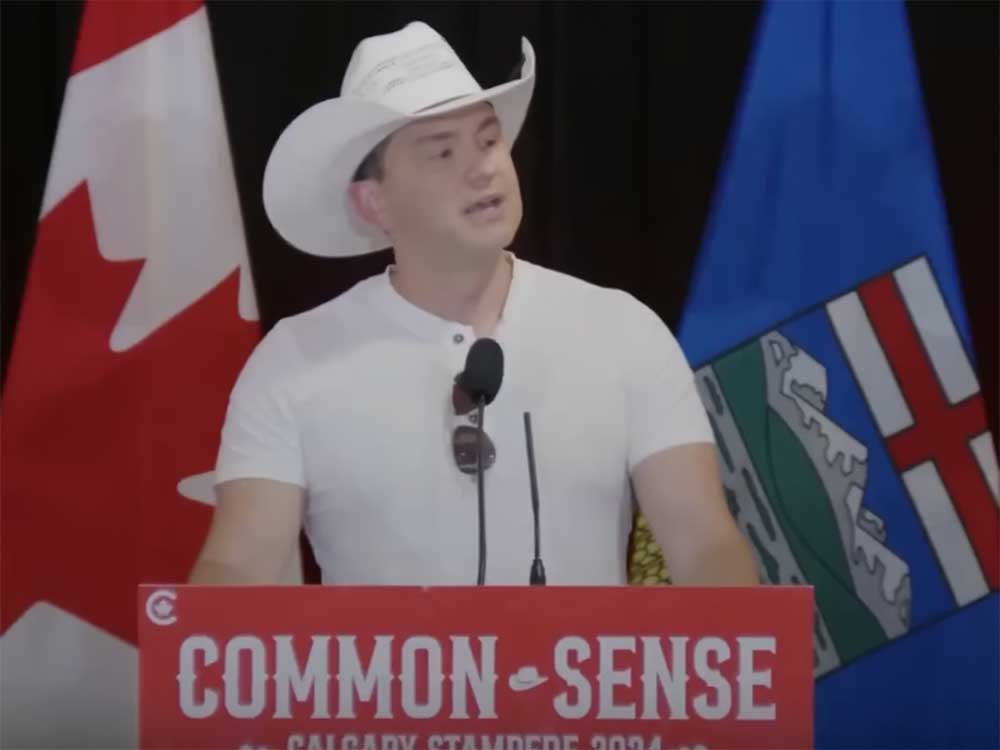 A 45-year-old light-skinned man with short dark hair wearing a white cowboy hat and T-shirt stands at a podium.