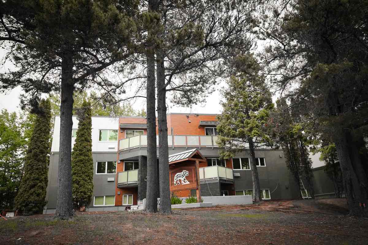 A three-storey building painted white, grey and orange stands among a stand of trees. Indigenous artwork of a bear is visible on a wood structure outside the building.