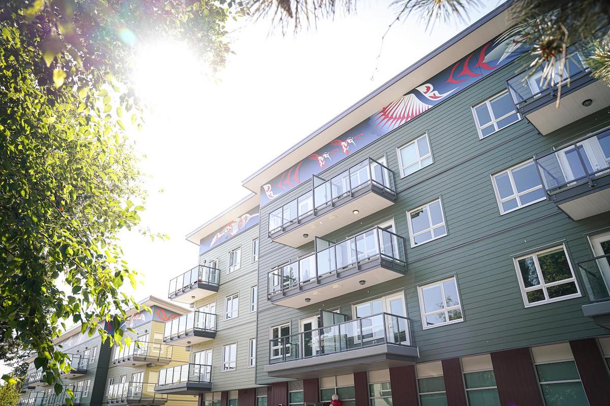 Sage-green low-rise apartment buildings shot from below feature the sun shining over them. Each building is decorated with Indigenous artwork in blue, red and white colourways on a horizontal plane across the top of each wall.