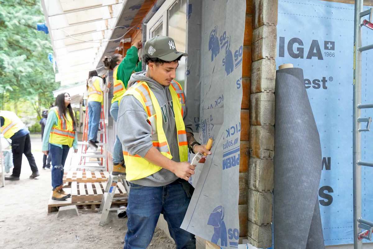 Young adults in bright yellow high-visibility vests over hoodies and jeans stand on ladders and steps to work on a residential building under construction. It’s a bright day and the wood walls are covered in a paper-like material.