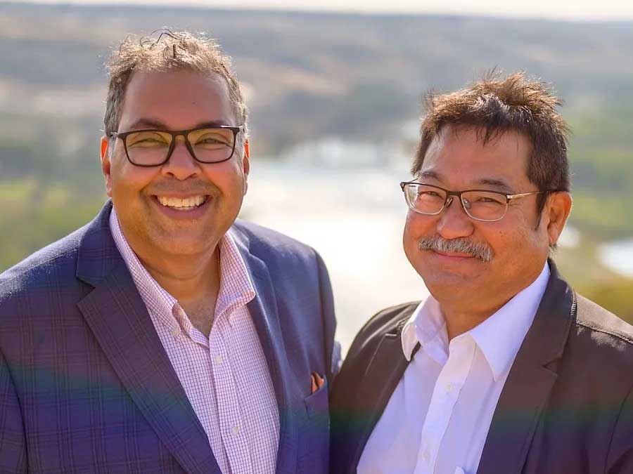 Two men, both with medium skin tones and wearing suit jackets with light-coloured shirts, smile at the camera.