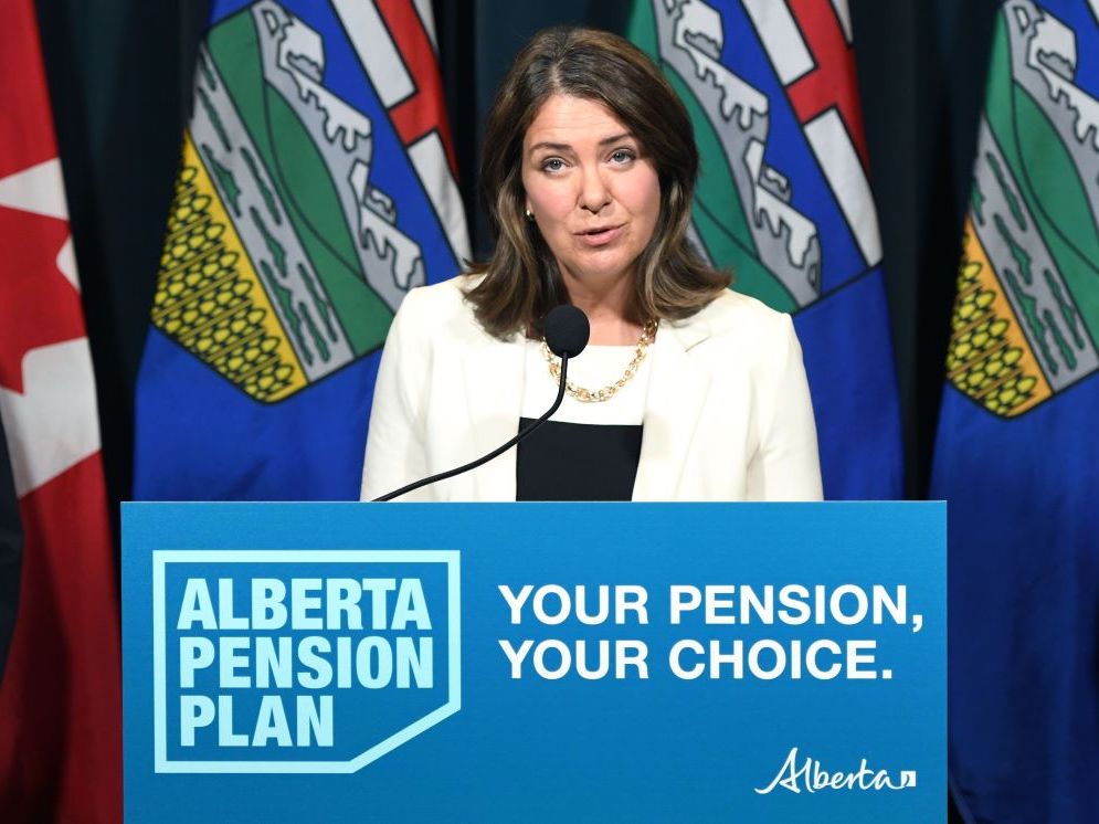 Danielle Smith, a light-skinned woman in her mid-50s with shoulder-length brown hair, wears a white blazer and gold necklace. She stands at a podium with a sign that says ‘Alberta Pension Plan, Your Pension, Your Choice.’