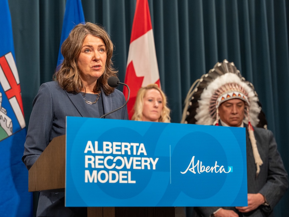 A 53-year-old woman with shoulder length brown hair stands at a podium which has a blue sign saying "Alberta Recovery Model."