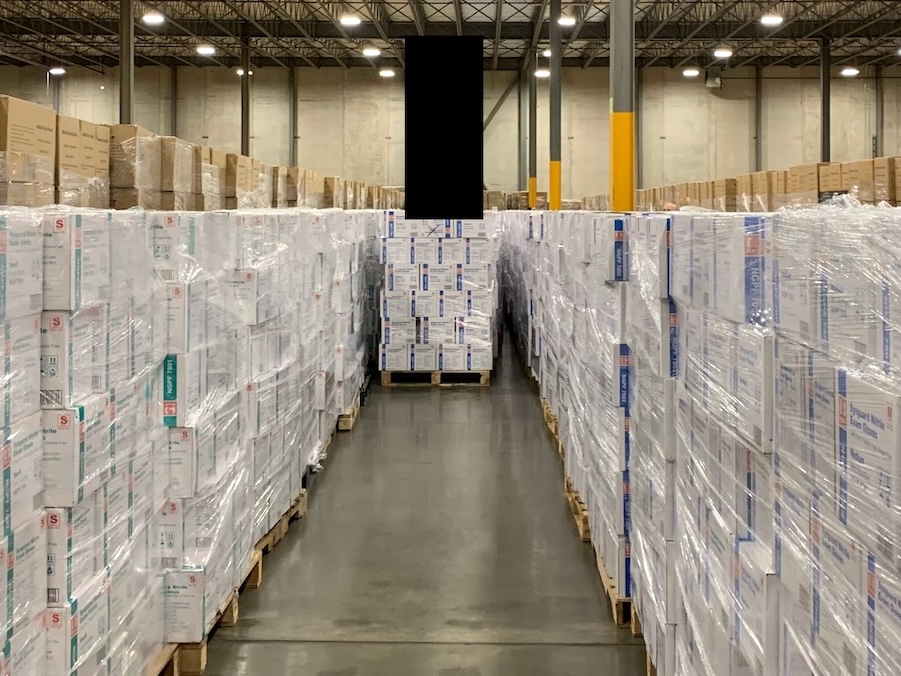 Pallets of shrink-wrapped white cardboard boxes are in a warehouse, surrounded by pallets of larger brown cardboard boxes.