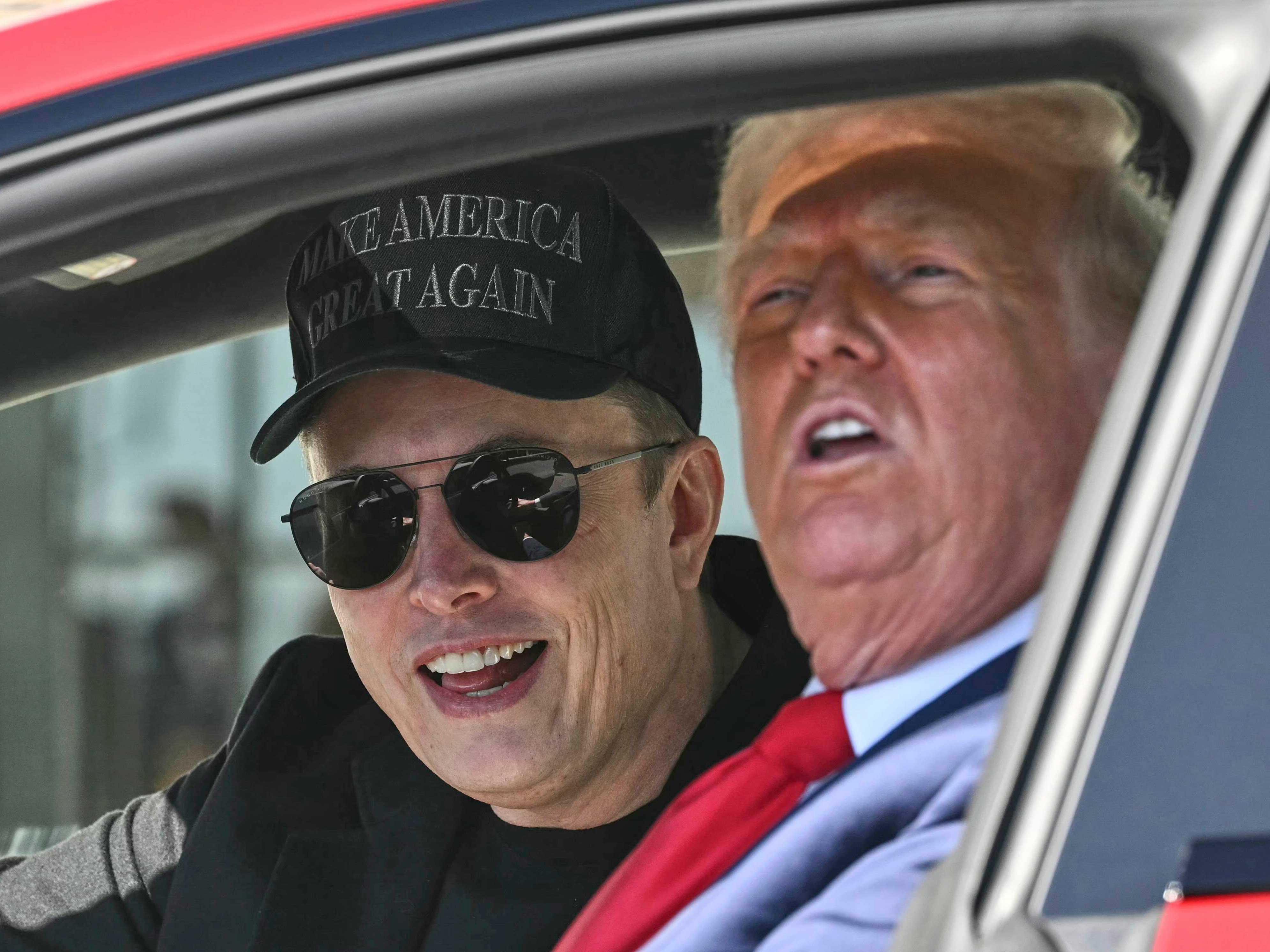 Elon Musk, wearing aviator sun glasses and a black MAGA hat, sits beside Donald Trump in a red Tesla.