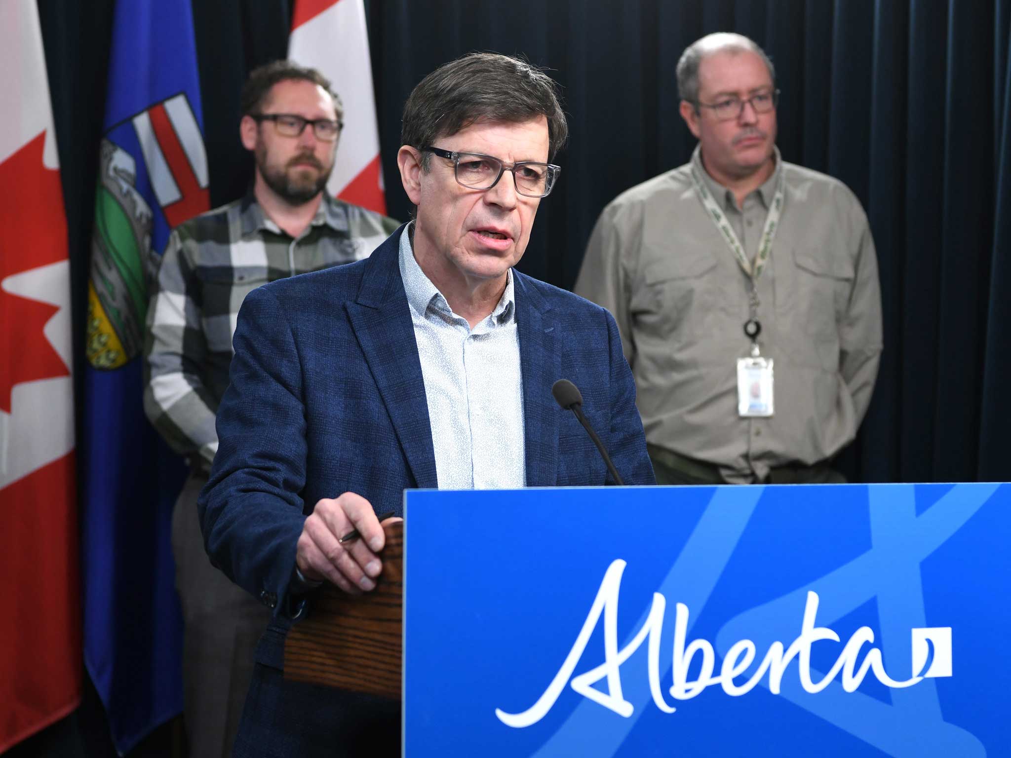 A 58-year-old light-skinned man stands at a podium, wearing a blue sports jacket and white shirt with no tie. Two other men look on.