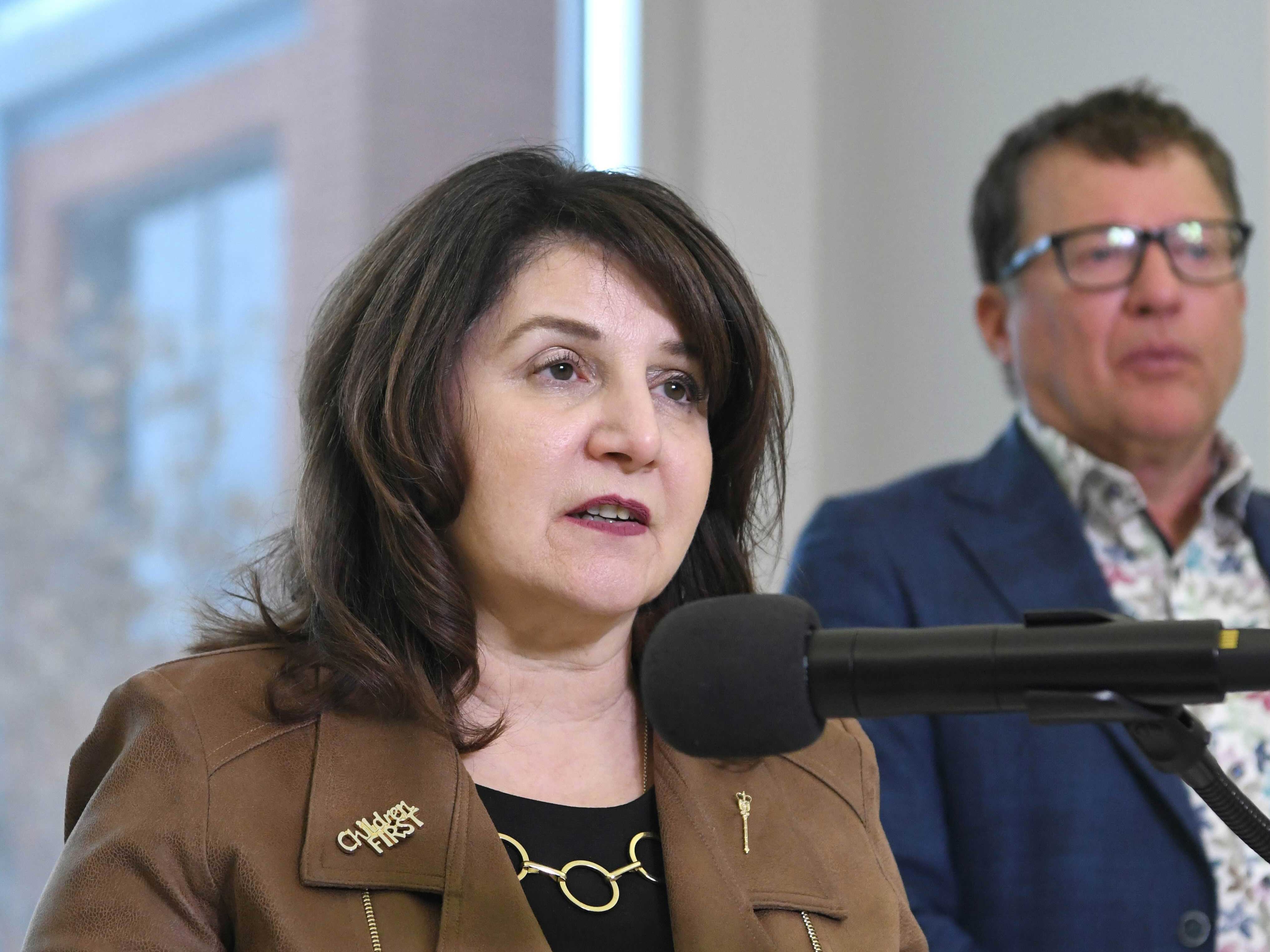 Adriana LaGrange, a light-skinned 64-year-old woman with brown hair, stands at a podium.