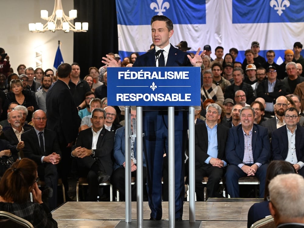 Pierre Poilievre has dark, slicked-back hair and pale skin. He is wearing a dark suit and standing at a podium with a blue sign with white text that reads ‘Fédéralisme responsable.’ A Quebec flag hangs in the background. He is in an indoor space and a large supportive audience is seated behind him.