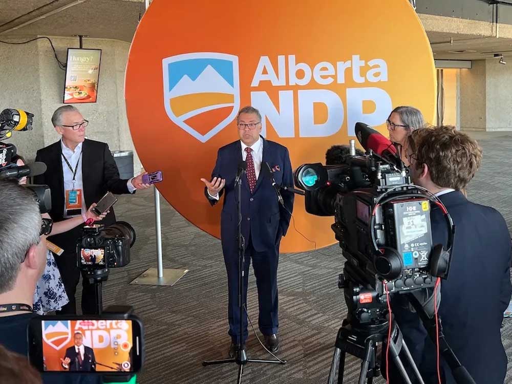 Naheed Nenshi, a middle-aged man with medium-skin tones, speaks to reporters in front of a large orange Alberta NDP logo.