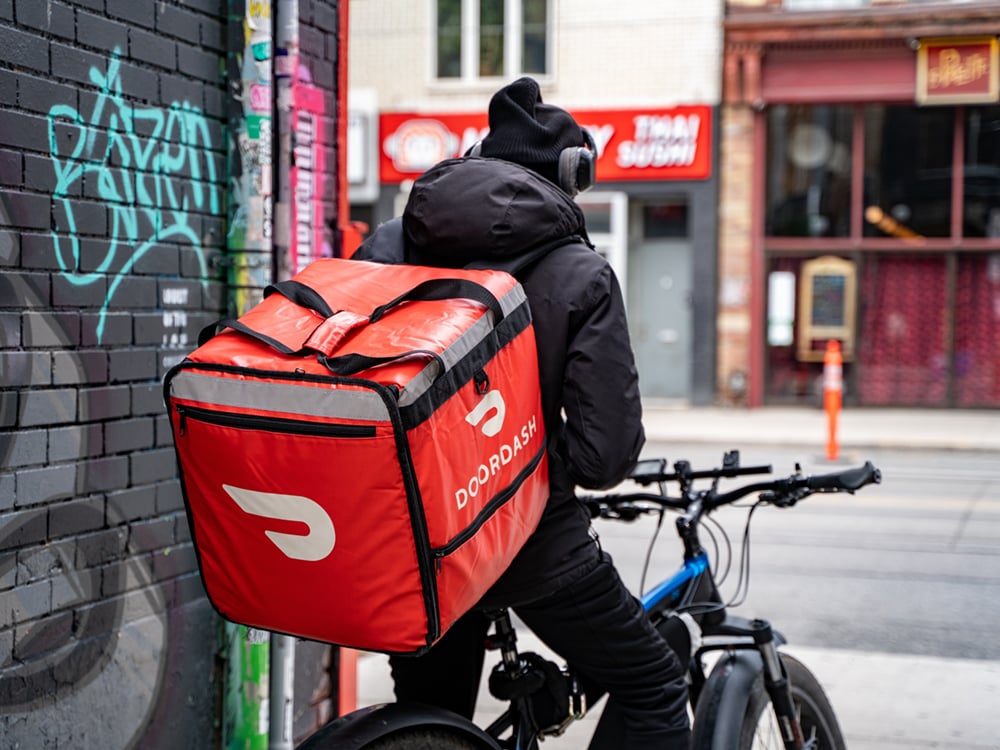 A person wears a large DoorDash backpack and rides an e-bike in the city. 