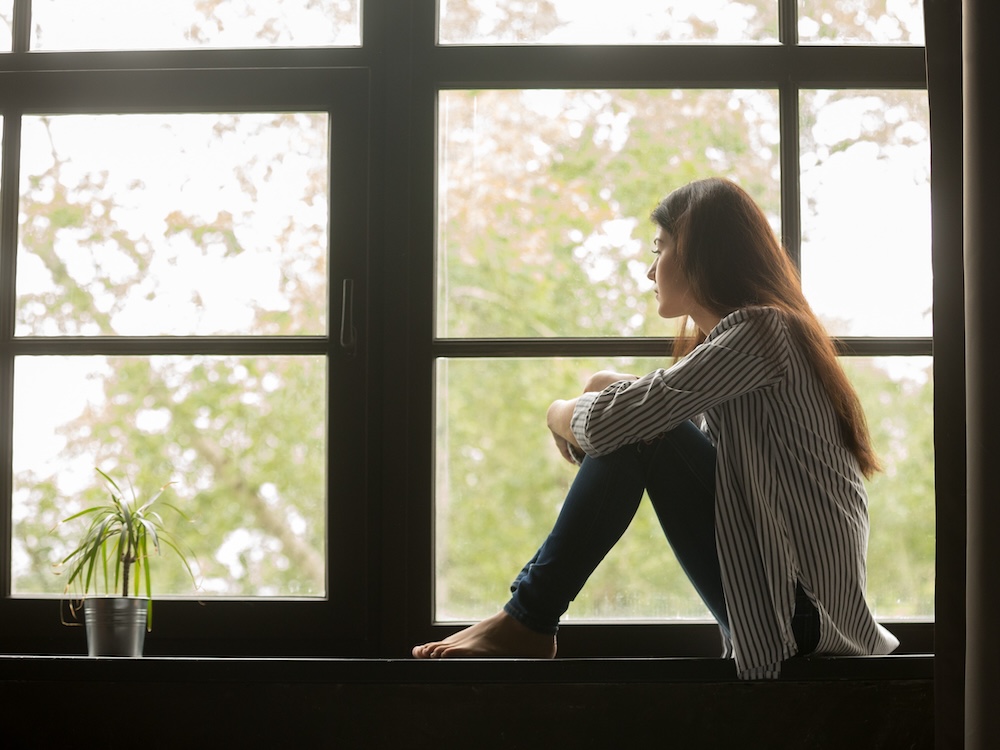 A wowan with black hair and Asian features looks out a window with blurred green outside.