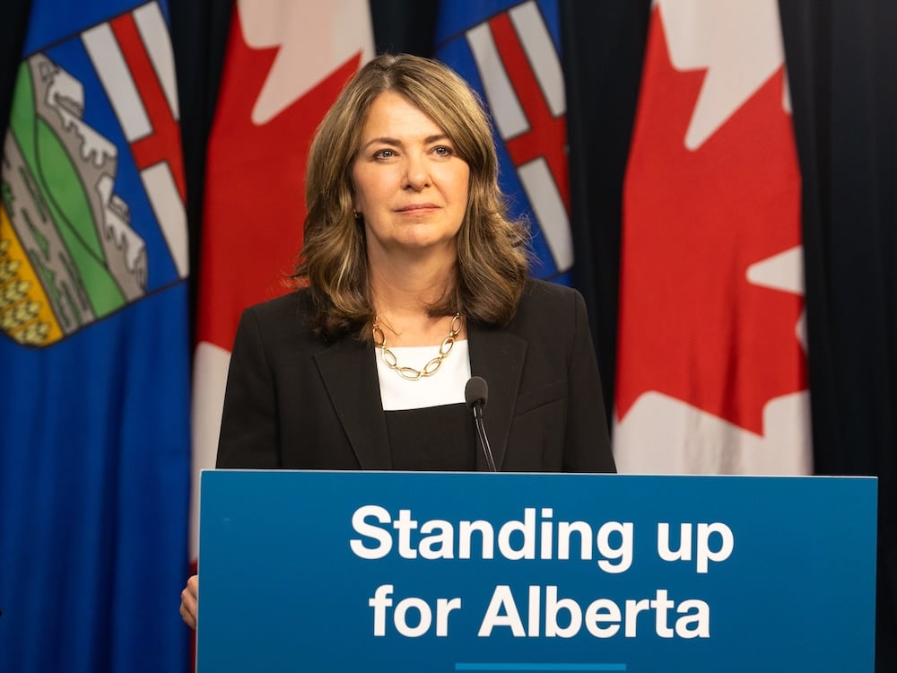 Danielle Smith, a 54-year-old light-skinned woman with shoulder-length brown hair, stands at a lectern with a sign saying, ‘Standing Up for Alberta.’