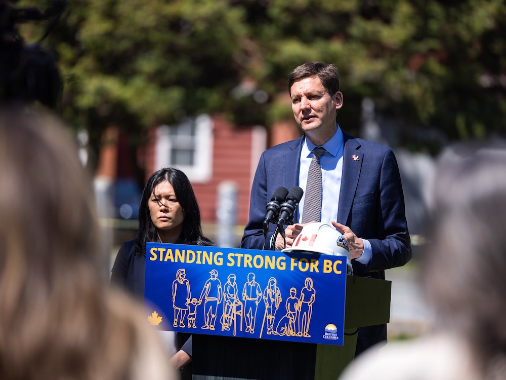 Bowinn Ma, a 39-year-old woman with medium skin tone, stands near David Eby, who is at a podium that says ‘Standing Strong for BC.’