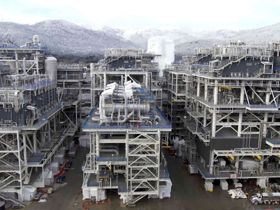 Giant grey industrial structures, with pipes and tanks, stand in front of snowy hills.