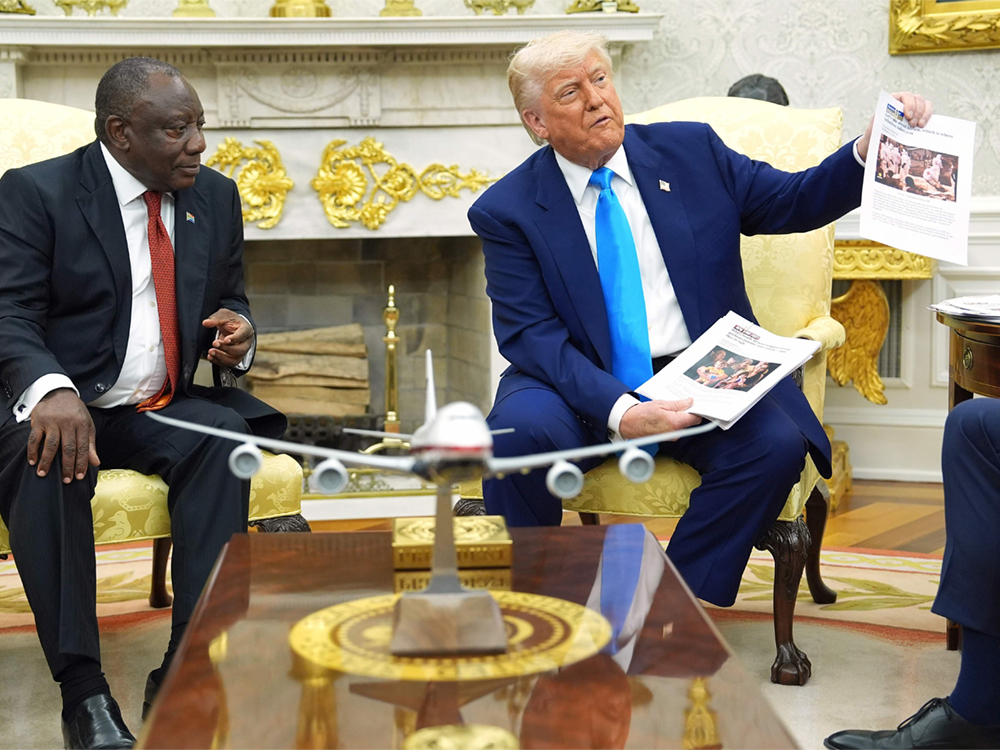 Cyril Ramaphosa, a 72-year-old Black man wearing a dark suit, sits in the Oval Office beside Donald Trump, who holds papers.