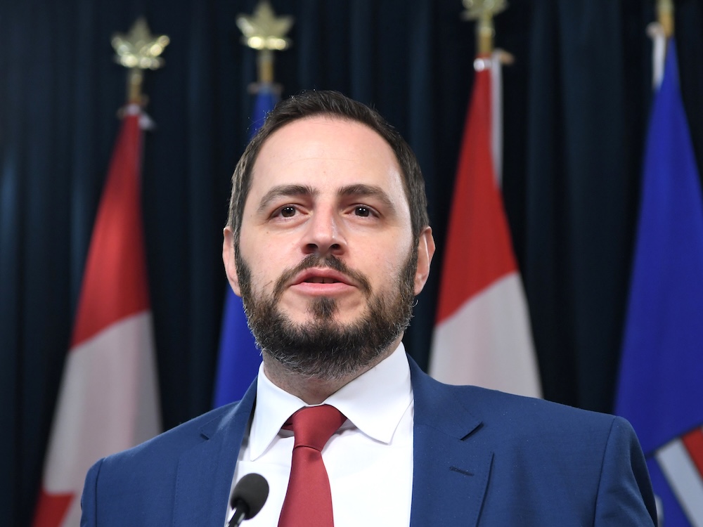 A 42-year-old light-skinned man with short dark hair and a beard wears a suit as he stands at a lectern.
