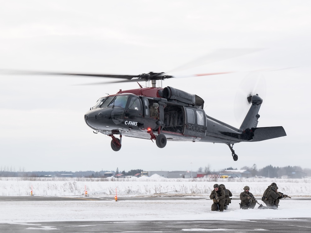 A black helicopter hovers above four armed people in military garb in a snowy landscape.