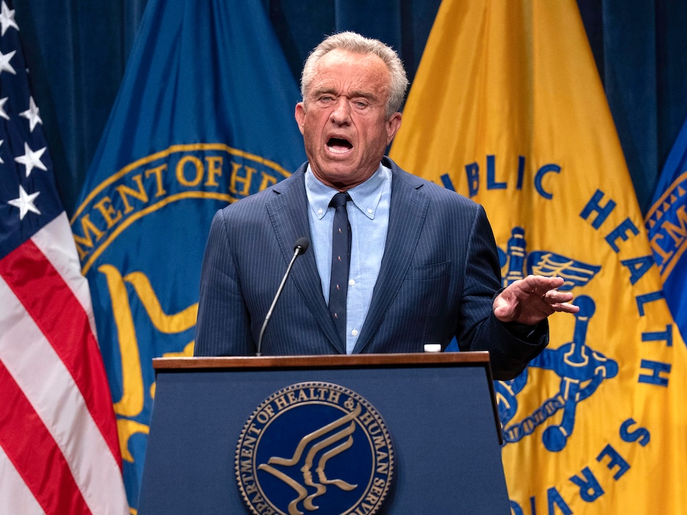 A 71-year-old man with tanned light skin and short grey hair, wearing a suit, speaks at a lectern that says &lsquo;Department of Health and Human Services.&rsquo;