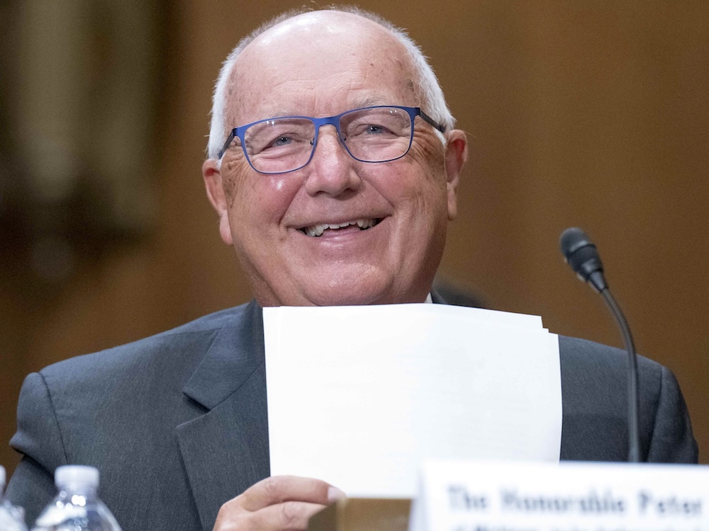 A 71-year-old light-skinned man with short white hair and blue-framed glasses smiles while holding papers.
