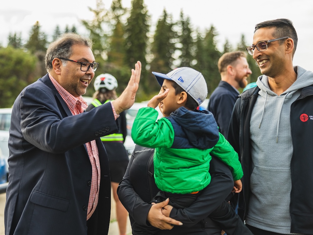 Naheed Nenshi, a burly man with medium-toned skin exchanges a high five with a child as others look on.