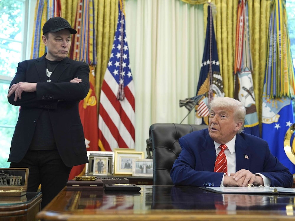 Elon Musk, in a black jacket and ball cap, stands looking down at Donald Trump, who is sitting at a desk.