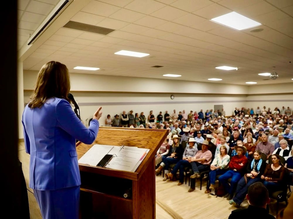Danielle Smith, in a pale blue pantsuit, is shown from behind as she addresses a large crowd in a gymnasium-like hall.