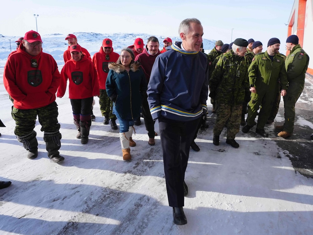Mark Carney has short greying dark hair and a light skin tone. He is wearing a blue top with yellow, blue and white trim over dark business clothing. He is surrounded to the left of the frame by members of the Canadian Rangers, in red ball caps and hoodies, and, to the right of the frame, by members of the Canadian Armed Forces in green fatigues.