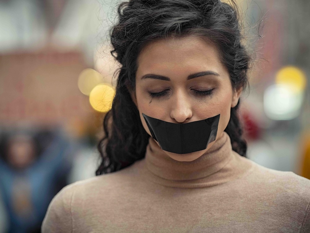 A light-skinned woman stands, eyes downcast, with a broad strip of black tape across her mouth.