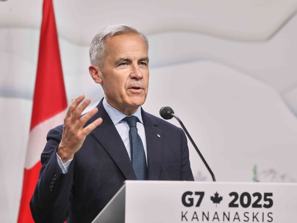 A 60-year-old light-skinned man with short hair and wearing a blue suit stands at a lectern with a sign that says ‘G7 2025 Kananaskis.’