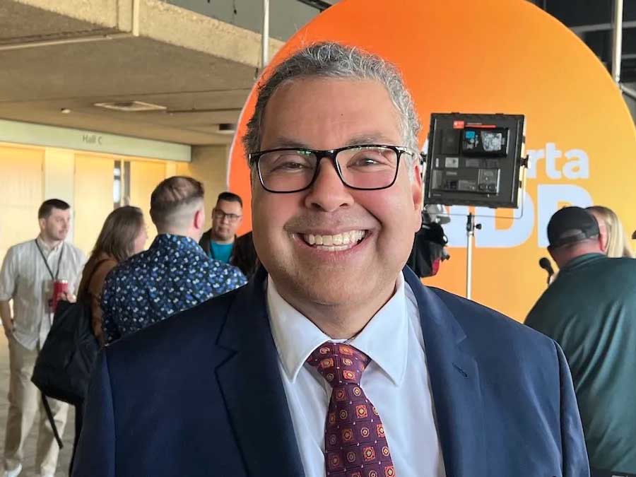 Naheed Nenshi, a 53-year-old man with medium-tone skin and glasses, grins. Behind him is a large round orange DP sign.