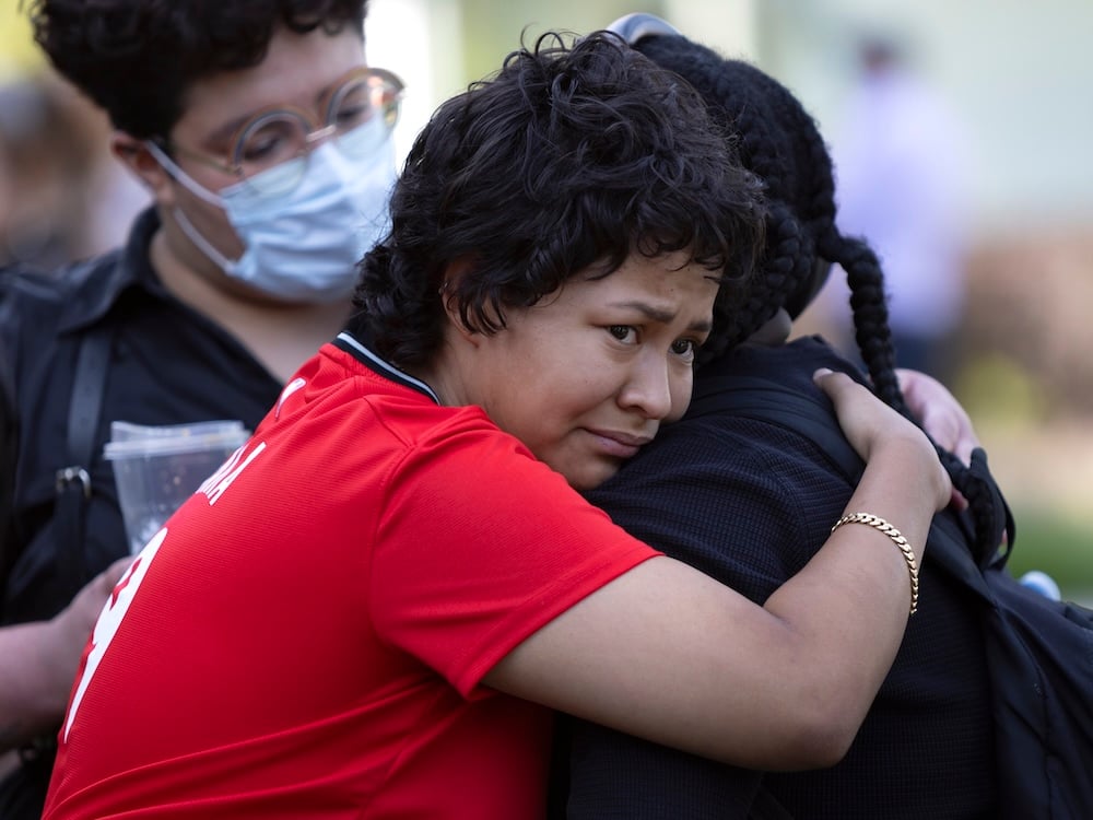 Two people embrace. The one facing the camera is a sad-looking person with medium-toned skin and short black hair, wearing a red T-shirt.