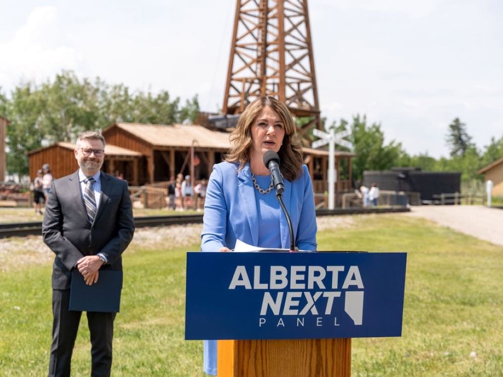 A 54-year-old light-skinned woman with shoulder-length brownish hair stands outdoors at a lectern that says ‘Alberta Next Panel.’ A 50-ish man with a grey beard, wearing a suit, stands behind.
