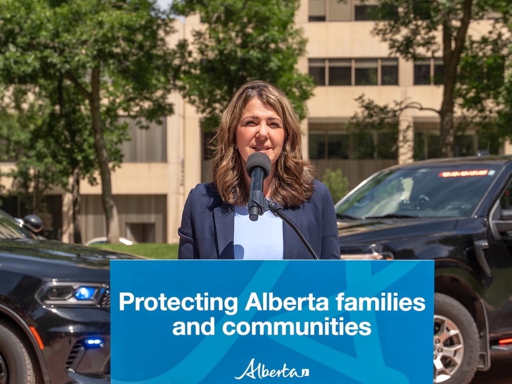 A 54-year-old light-skinned woman with long dark hair wears a blue suit and stands outdoors at a podium that says ‘Protecting Alberta families and communities.’