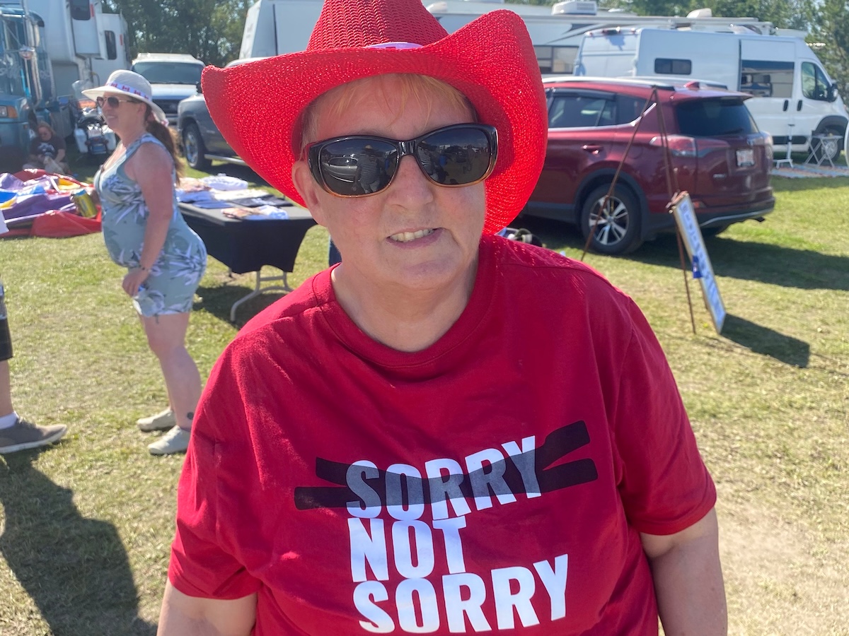 A 60-ish woman is wearing a red cowboy hat, sunglasses and a red T-shirt that says ‘Sorry, Not Sorry,’ with the first ‘Sorry’ crossed out.