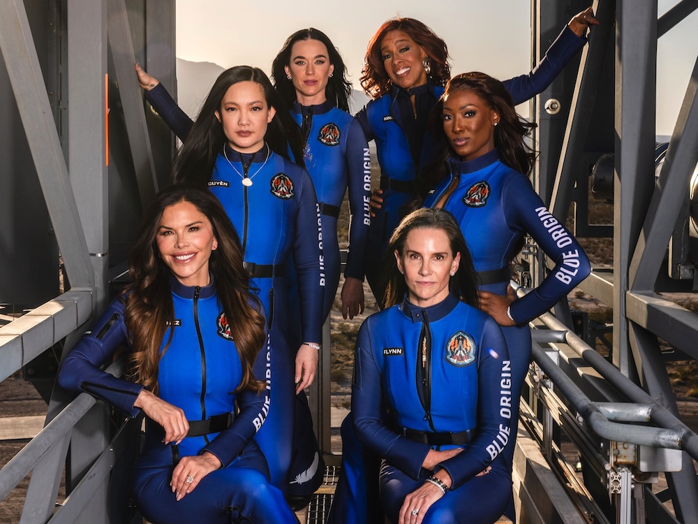 Six women of varying skin tones pose in flashy blue suits in front of a metal structure.