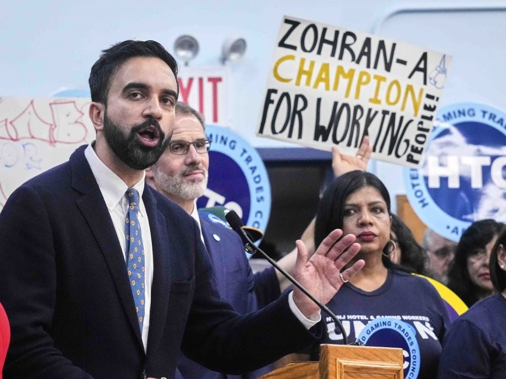 Zohran Mamdani has short dark hair and a beard. He is wearing a dark suit over a white shirt and speaking into a microphone at a wooden lectern during a rally. Behind him, people are holding signs including a handwritten one that reads ‘Zohran — a champion for working people.’