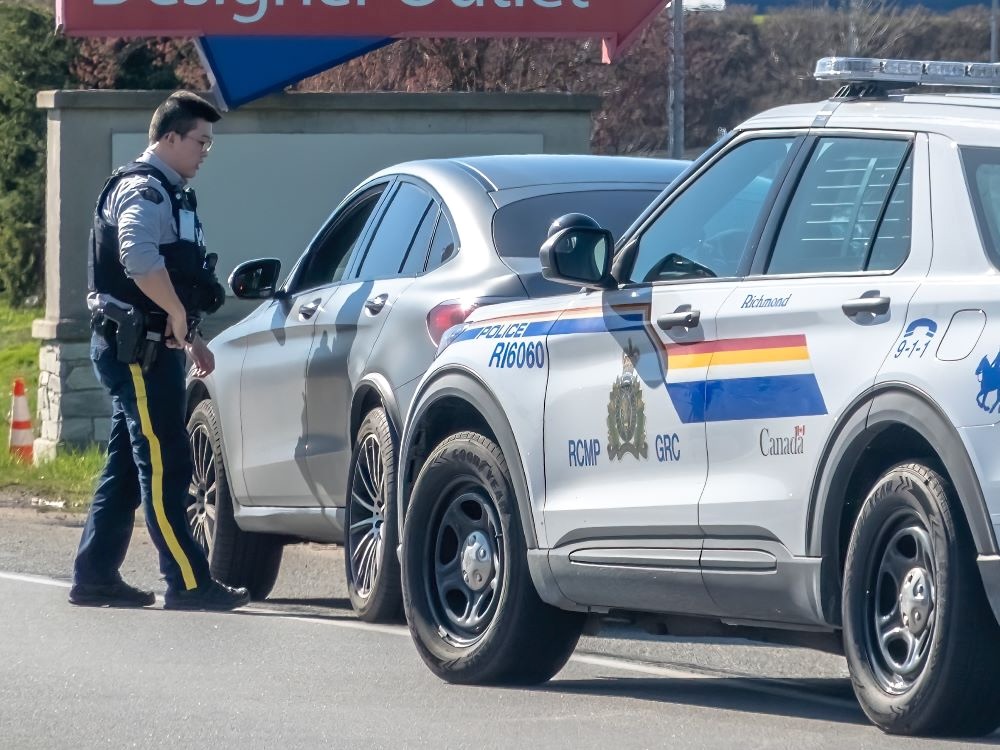 An RCMP officer stands next to a car speaking through the driver&rsquo;s-side open window parked streetside. Behind that car is parked an RCMP police car.