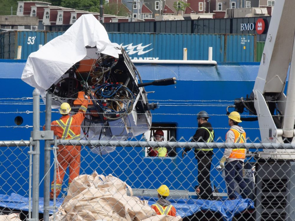 A chunk of wreckage, partly covered with a white tarp, is being unloaded from a ship by workers in hard hats and safety clothes.