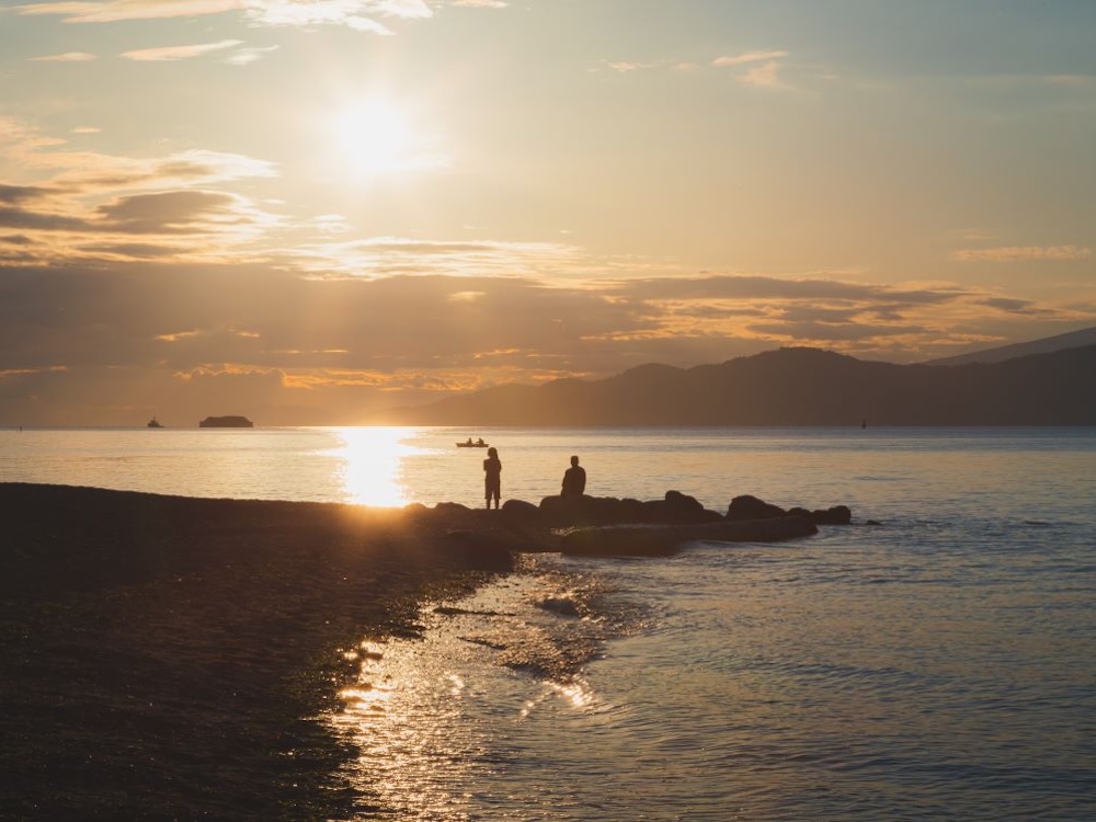 The sun sets over the sea, with people silhouetted on a rocky shore.